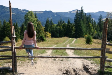 Young woman sitting on wooden fence and enjoying mountain landscape, back view. Space for text