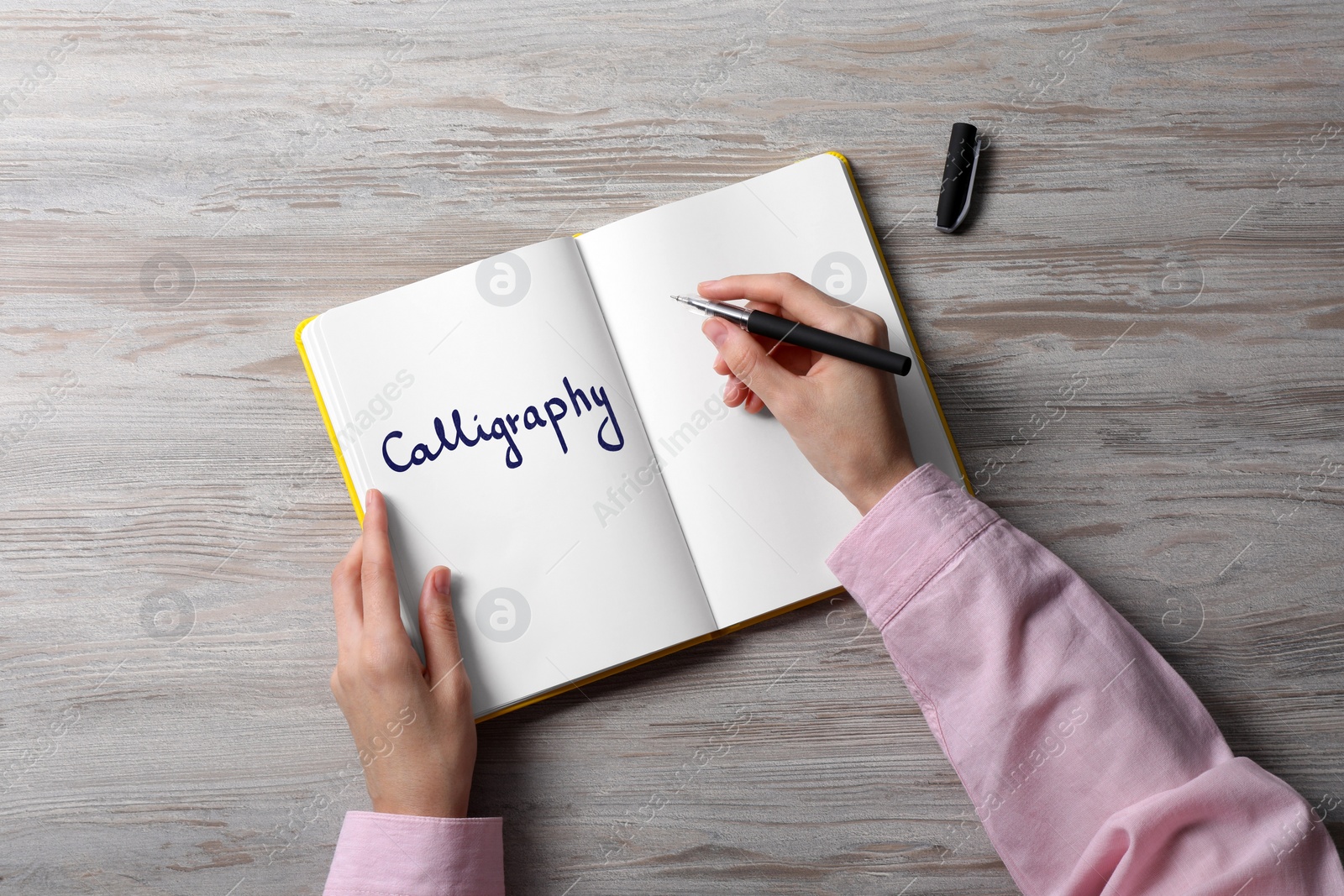 Image of Woman writing word Calligraphy in notebook at white wooden table, top view