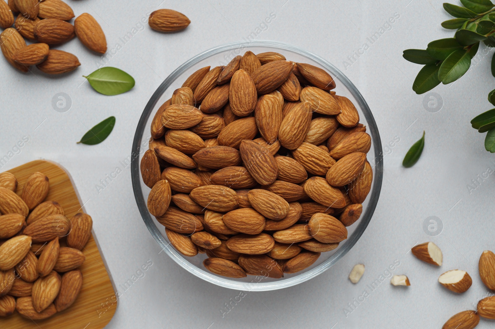 Photo of Bowl of delicious almonds and fresh leaves on white tiled table, flat lay