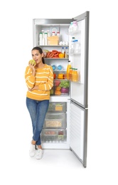 Photo of Young woman eating apple near open refrigerator on white background