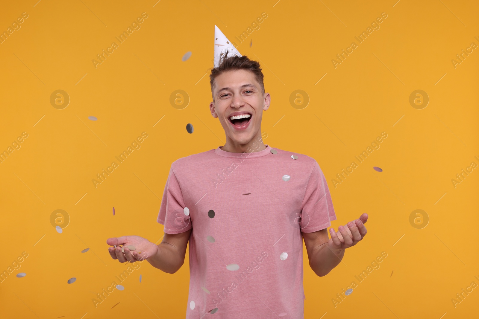 Photo of Happy man in party hat throwing confetti on orange background