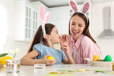 Happy mother and daughter with bunny ears headbands having fun while painting Easter egg in kitchen