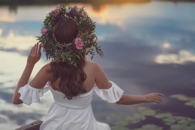 Young woman wearing wreath made of beautiful flowers near river in evening, back view