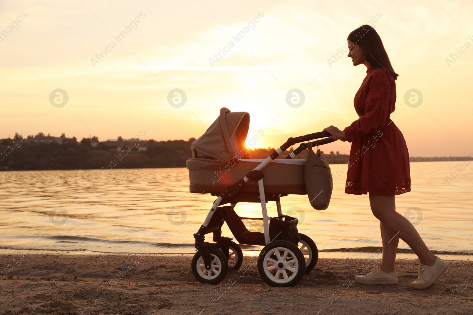 Photo of Happy mother with baby in stroller walking near river at sunset