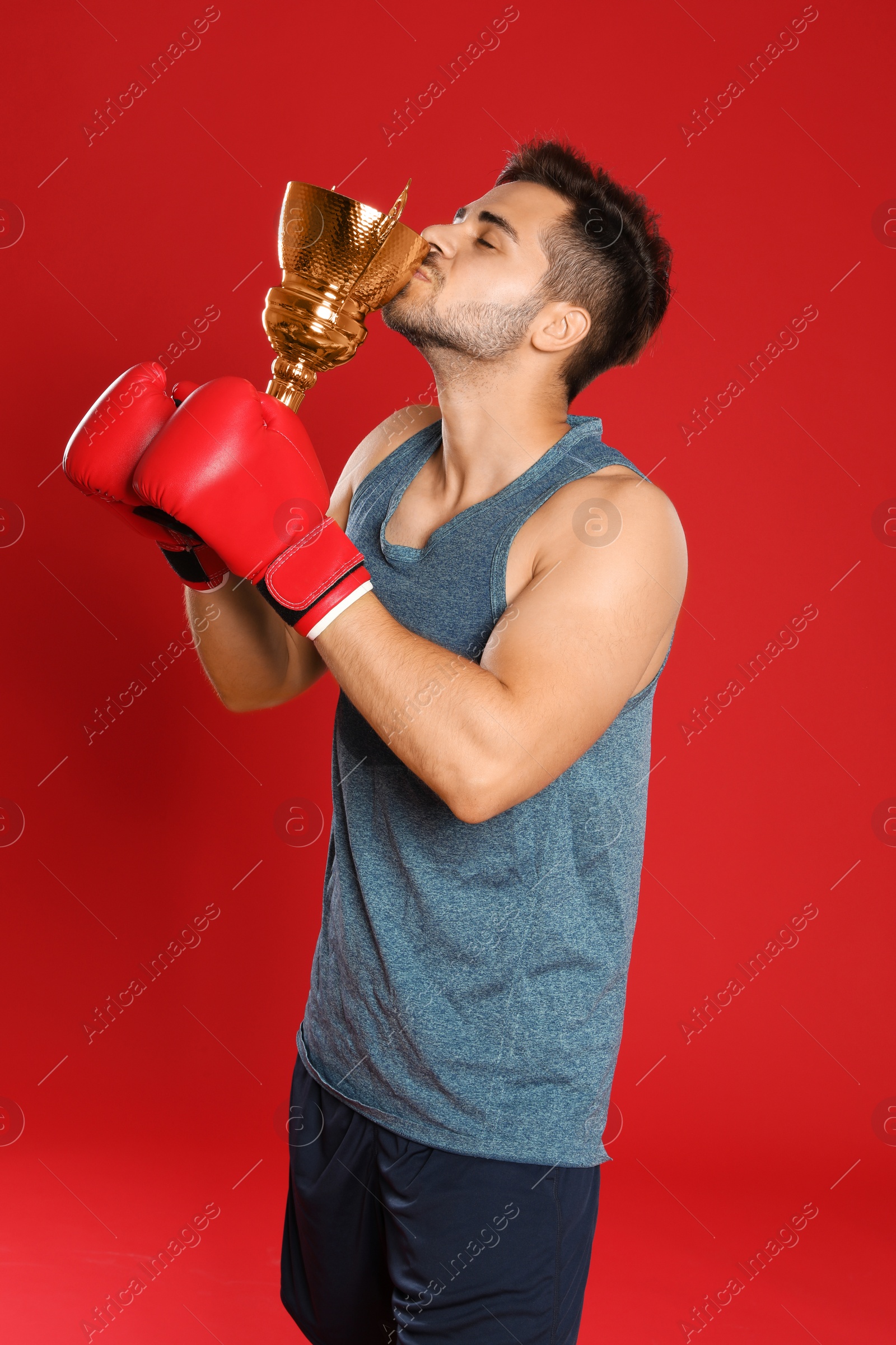 Photo of Portrait of young boxer kissing gold trophy cup on red background