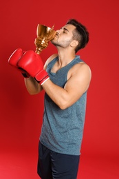 Portrait of young boxer kissing gold trophy cup on red background