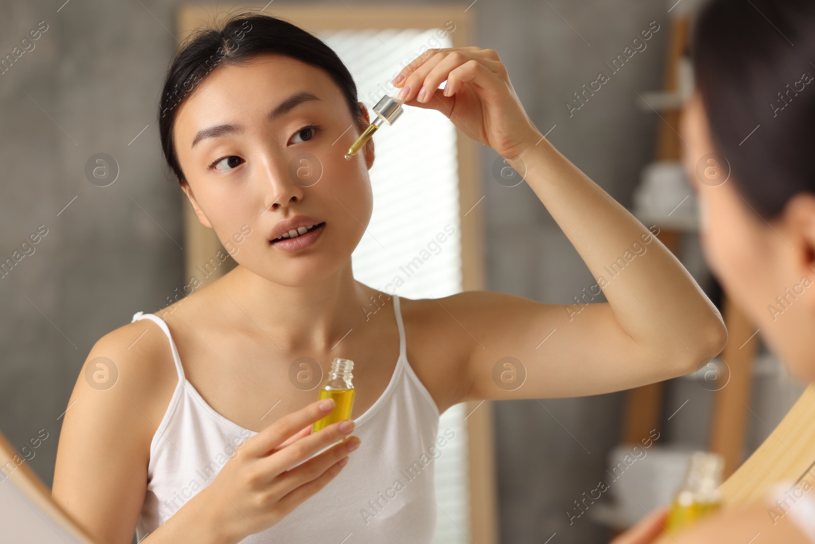 Photo of Beautiful young woman applying cosmetic serum onto her face near mirror in bathroom