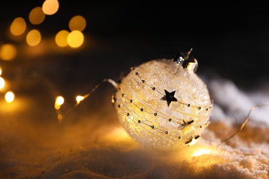 Photo of Christmas ball and fairy lights on white snow outdoors. Space for text