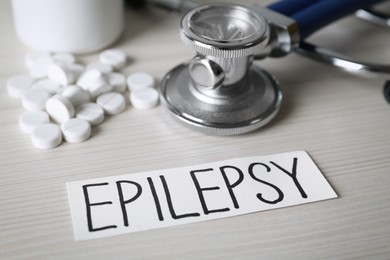 Photo of Card with word Epilepsy, stethoscope and pills on white wooden table, closeup