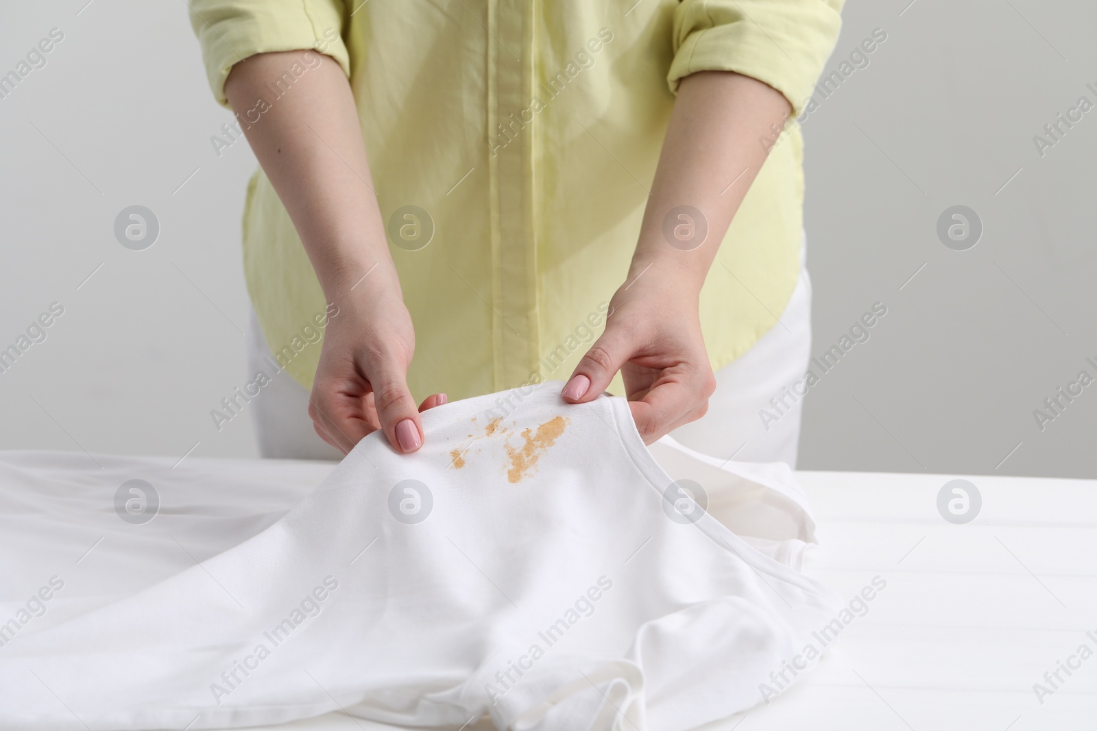 Photo of Woman holding shirt with stain at white table against light grey background, closeup