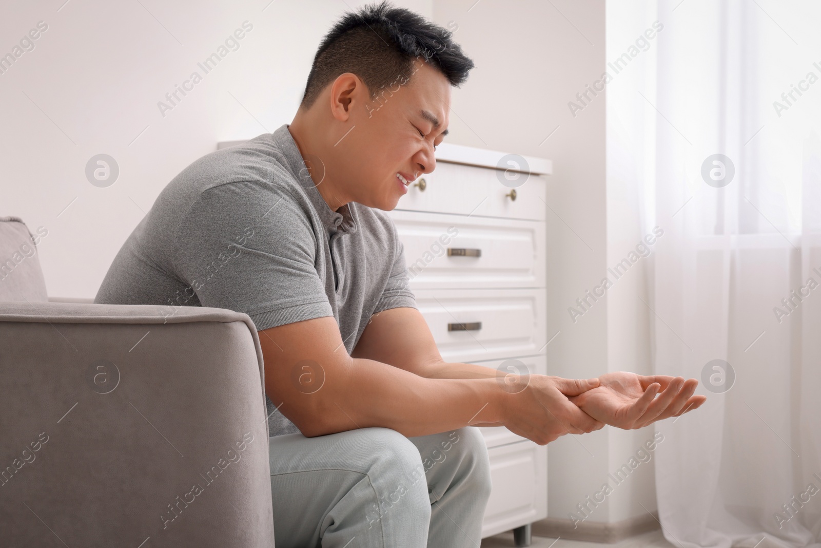 Photo of Asian man suffering from pain in his hand on armchair indoors