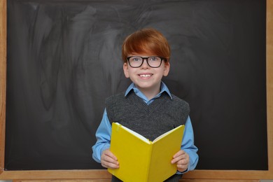 Smiling schoolboy in glasses with book near blackboard