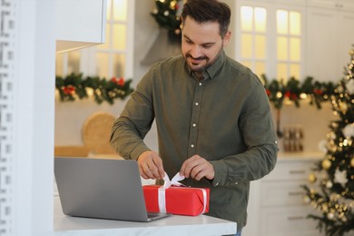 Photo of Celebrating Christmas online with exchanged by mail presents. Man opening gift box during video call on laptop in kitchen