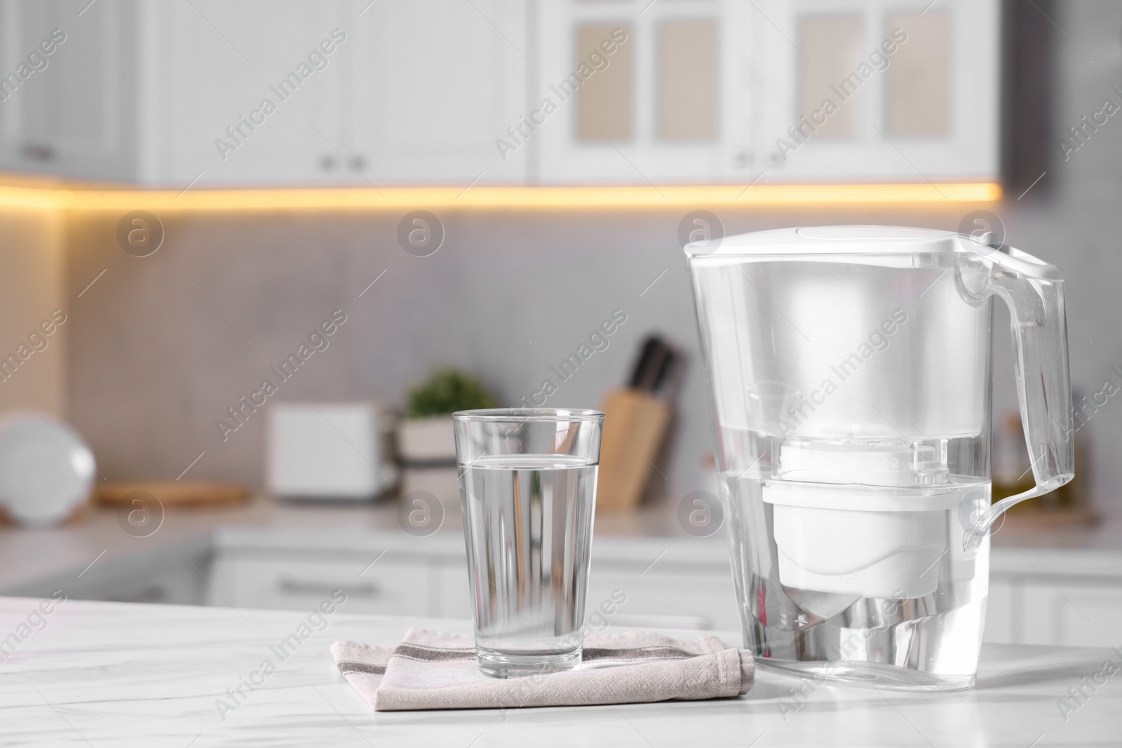 Photo of Water filter jug and glass on white marble table in kitchen, closeup. Space for text