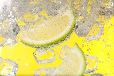 Photo of Juicy lime slices and ice cubes in soda water against yellow background, closeup