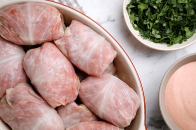 Uncooked stuffed cabbage rolls in baking dish on white marble table, flat lay