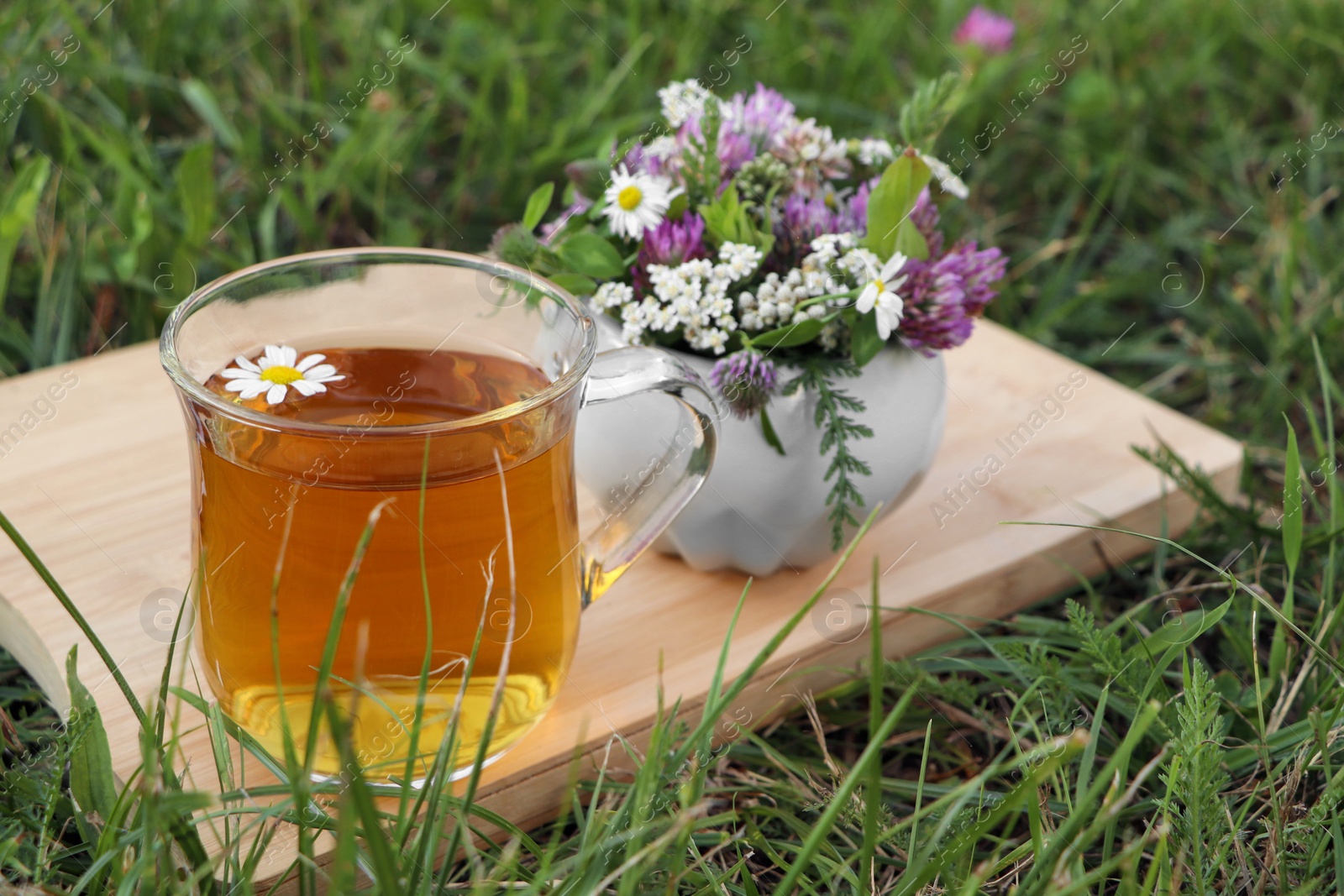 Photo of Cup of aromatic herbal tea and ceramic mortar with different wildflowers on green grass outdoors. Space for text