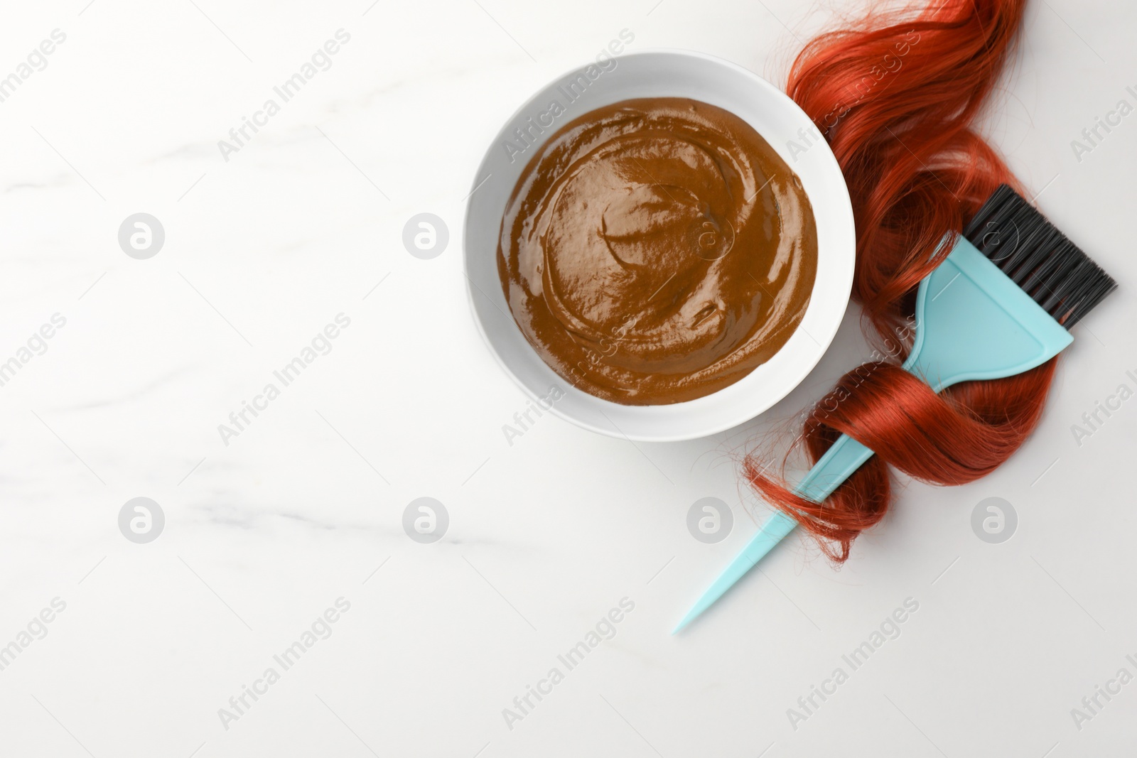 Photo of Bowl of henna cream, brush and red strand on white marble table, flat lay with space for text. Natural hair coloring