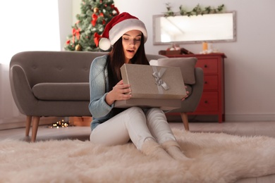 Beautiful young woman in Santa hat opening Christmas gift box at home