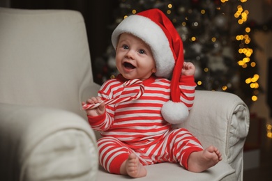Cute baby in Santa hat and bright Christmas pajamas holding candy cane at home