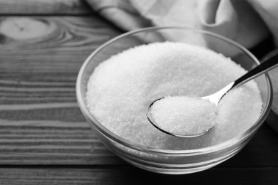 Photo of Granulated sugar in bowl and spoon on black wooden table, closeup. Space for text