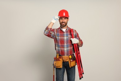 Photo of Professional builder in hard hat with tool belt and step ladder on light background