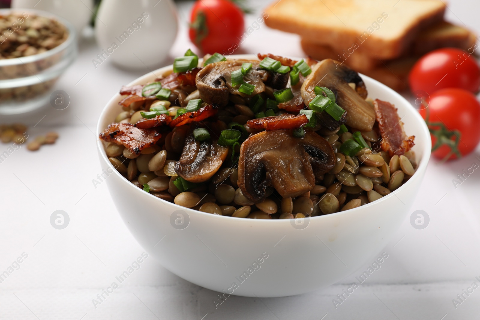 Photo of Delicious lentils with mushrooms, bacon and green onion in bowl served on white table, closeup