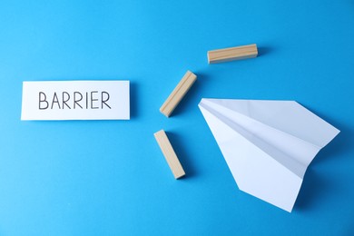 Photo of Paper plane movement blocked by wooden blocks and card with word Barrier on light blue background, flat lay. Development through obstacles overcoming