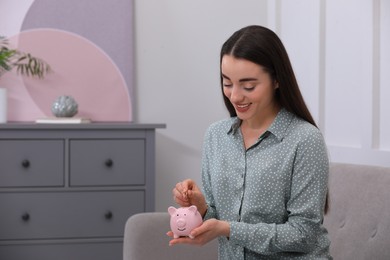 Photo of Young woman putting coin into piggy bank at home