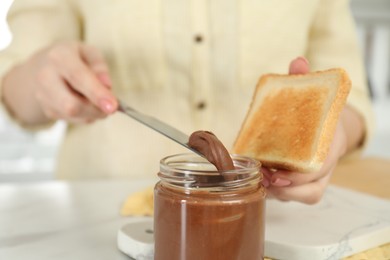 Photo of Woman with toast taking tasty nut butter onto knife at table, closeup