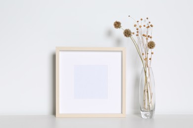 Photo of Empty photo frame and vase with dry decorative flowers on white table. Mockup for design