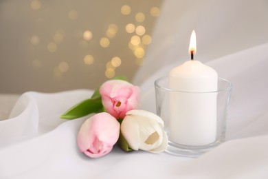 Photo of Burning candle in holder and beautiful tulips on white fabric against blurred lights, closeup