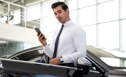 Photo of Young man with phone near car in modern dealership