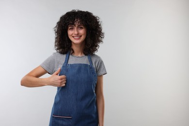 Photo of Happy woman wearing kitchen apron and showing thumbs up on light grey background, space for text. Mockup for design