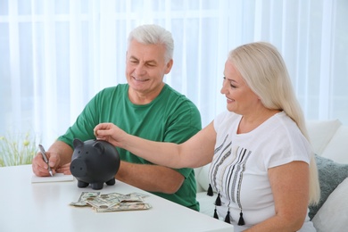 Photo of Mature woman putting money into piggy bank and her husband at table