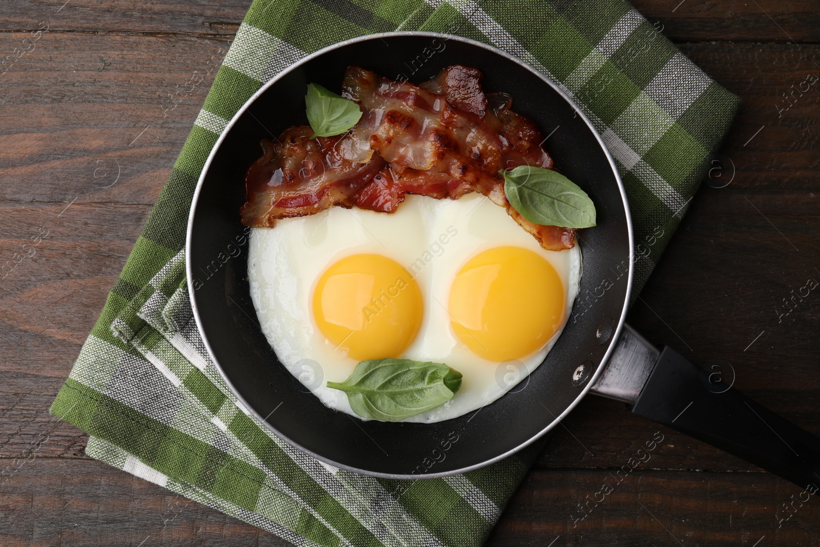 Photo of Fried eggs, bacon and basil in frying pan on wooden table, top view