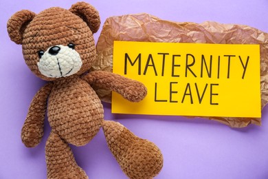 Photo of Toy bear and note with text Maternity Leave on violet background, flat lay