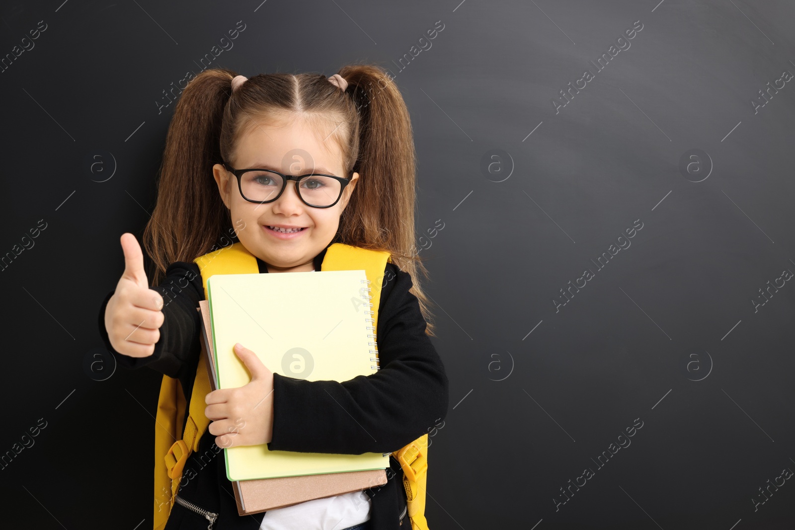 Photo of Happy little school child with notebooks showing thumbs up near chalkboard. Space for text