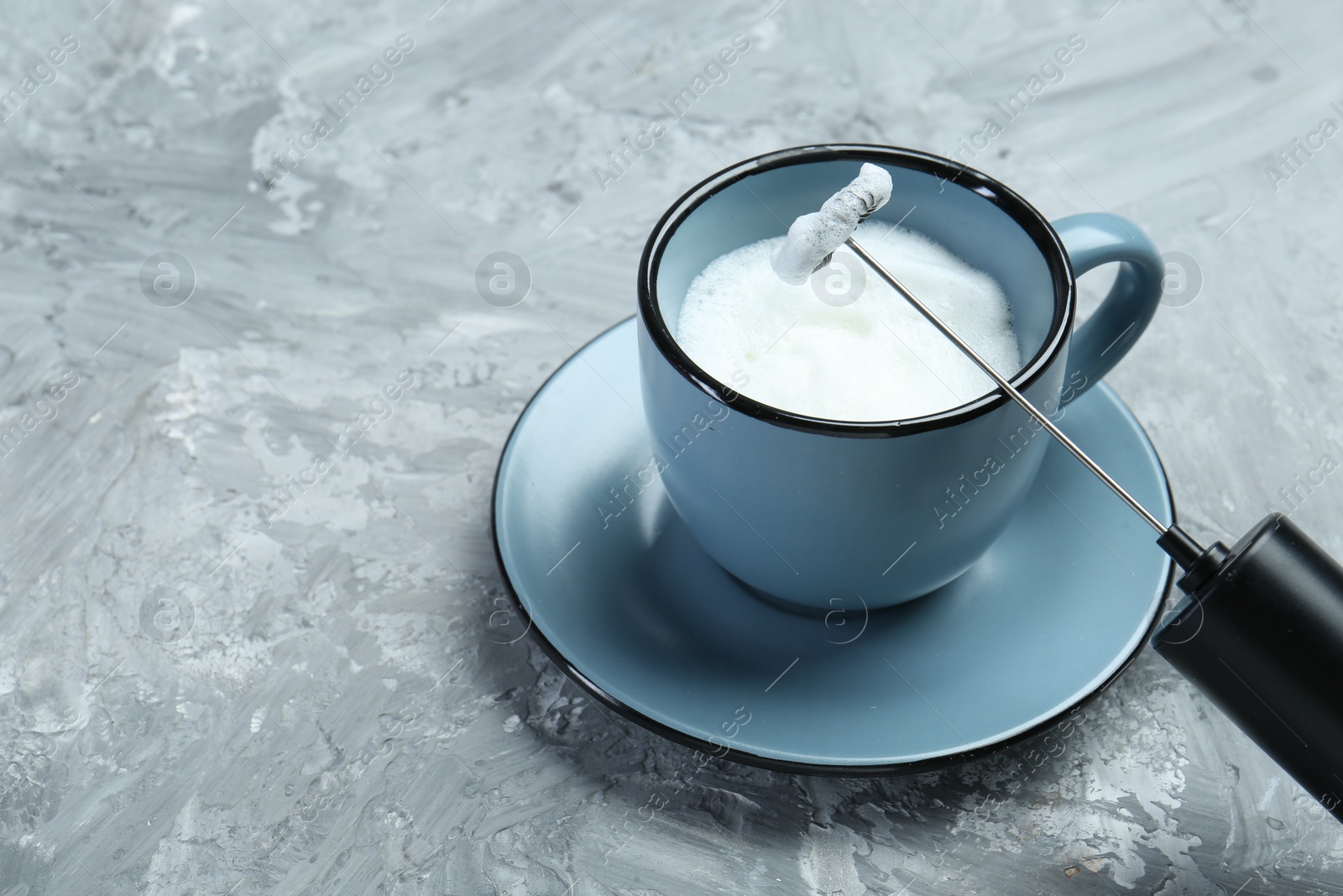 Photo of Mini mixer (milk frother) and whipped milk in cup on grey textured table, closeup. Space for text