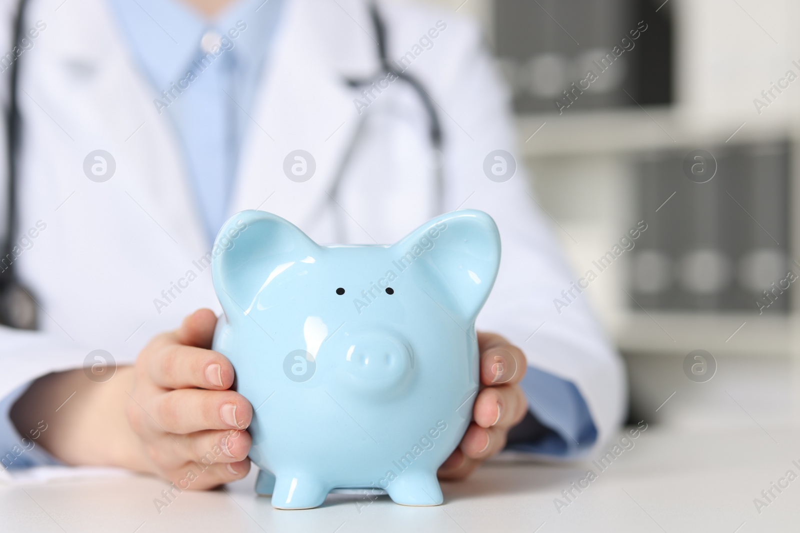 Photo of Doctor with piggy bank at white table indoors, closeup. Space for text