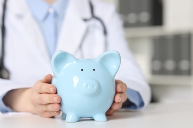 Photo of Doctor with piggy bank at white table indoors, closeup. Space for text