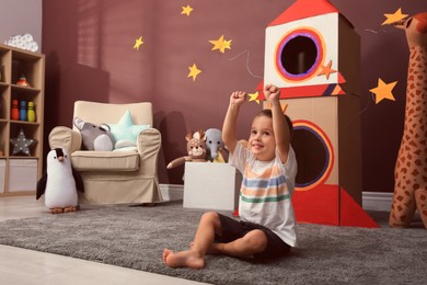 Photo of Cute little boy playing on floor near cardboard rocket  at home. Child's room interior