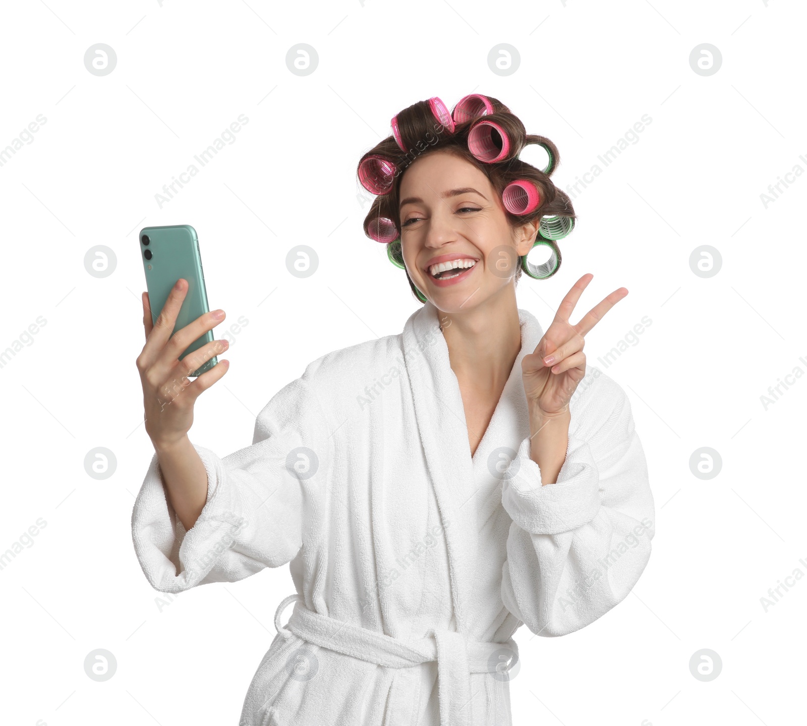 Photo of Happy young woman in bathrobe with hair curlers taking selfie on white background