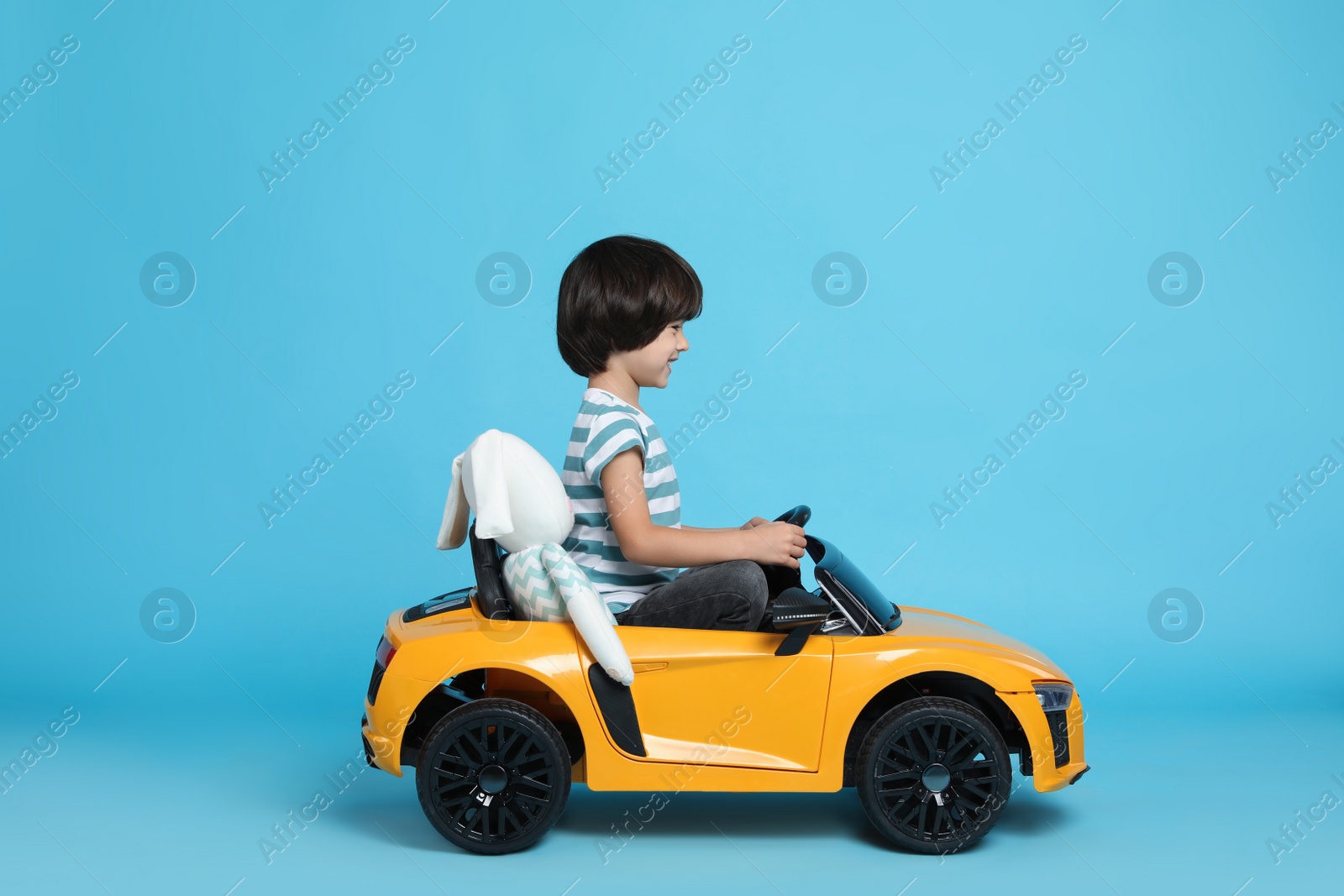 Photo of Cute little boy with toy bunny driving children's car on light blue background