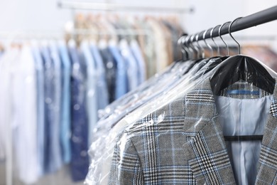 Photo of Dry-cleaning service. Many different clothes in plastic bags hanging on rack indoors, closeup