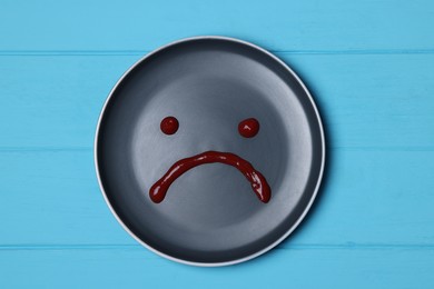 Photo of Sad face drawn on plate with ketchup on light blue wooden table, top view