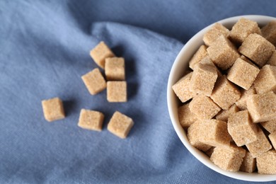 Photo of Brown sugar cubes in bowl on blue tablecloth, top view. Space for text