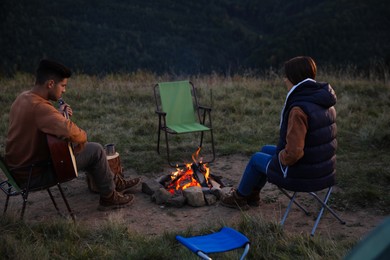 Photo of Couple with guitar sitting near bonfire at camping site