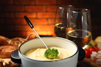 Photo of Fork with piece of broccoli, melted cheese in fondue pot, wine and snacks on blurred background, closeup