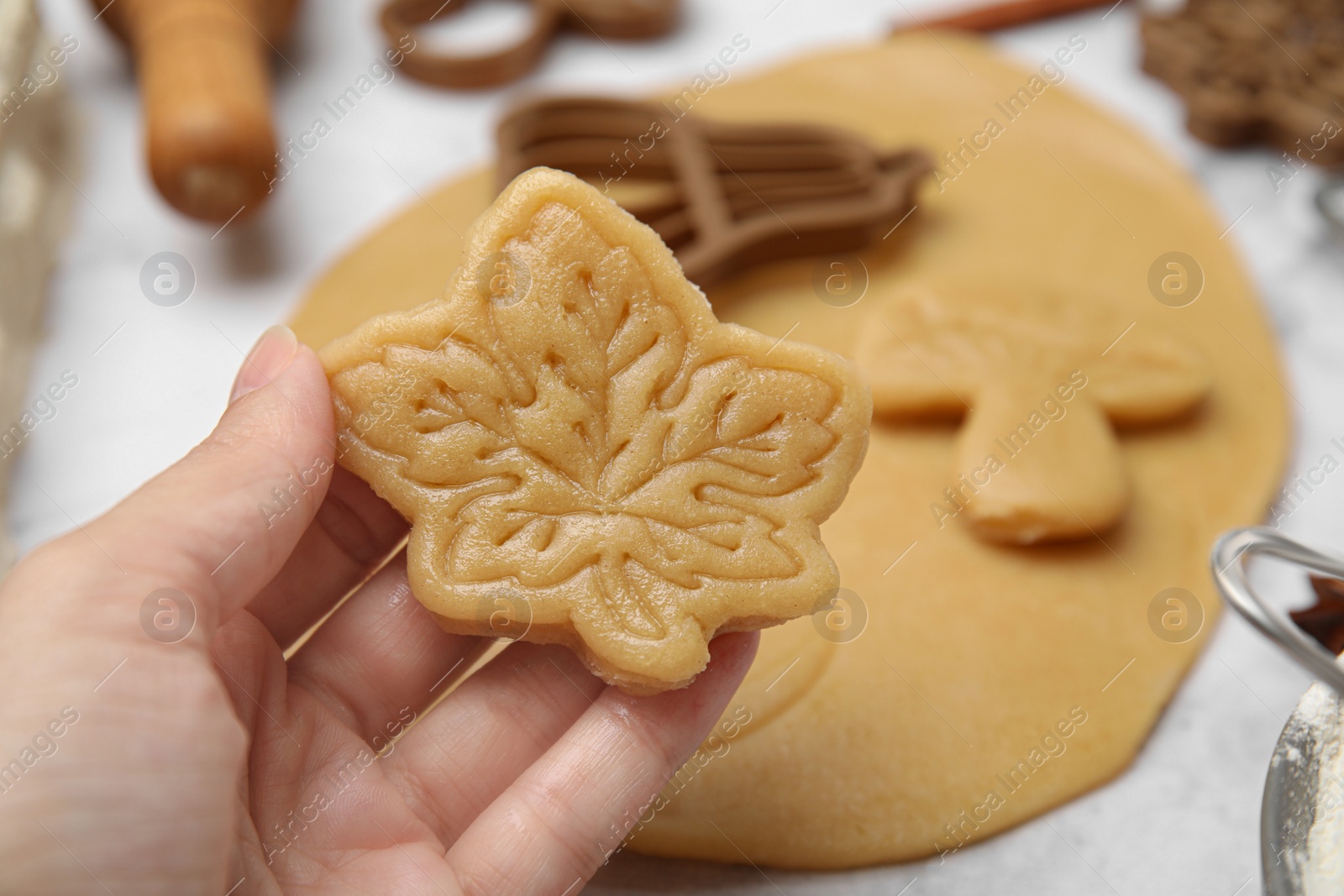 Photo of Woman holding unbaked leaf shaped cookie at white table, closeup